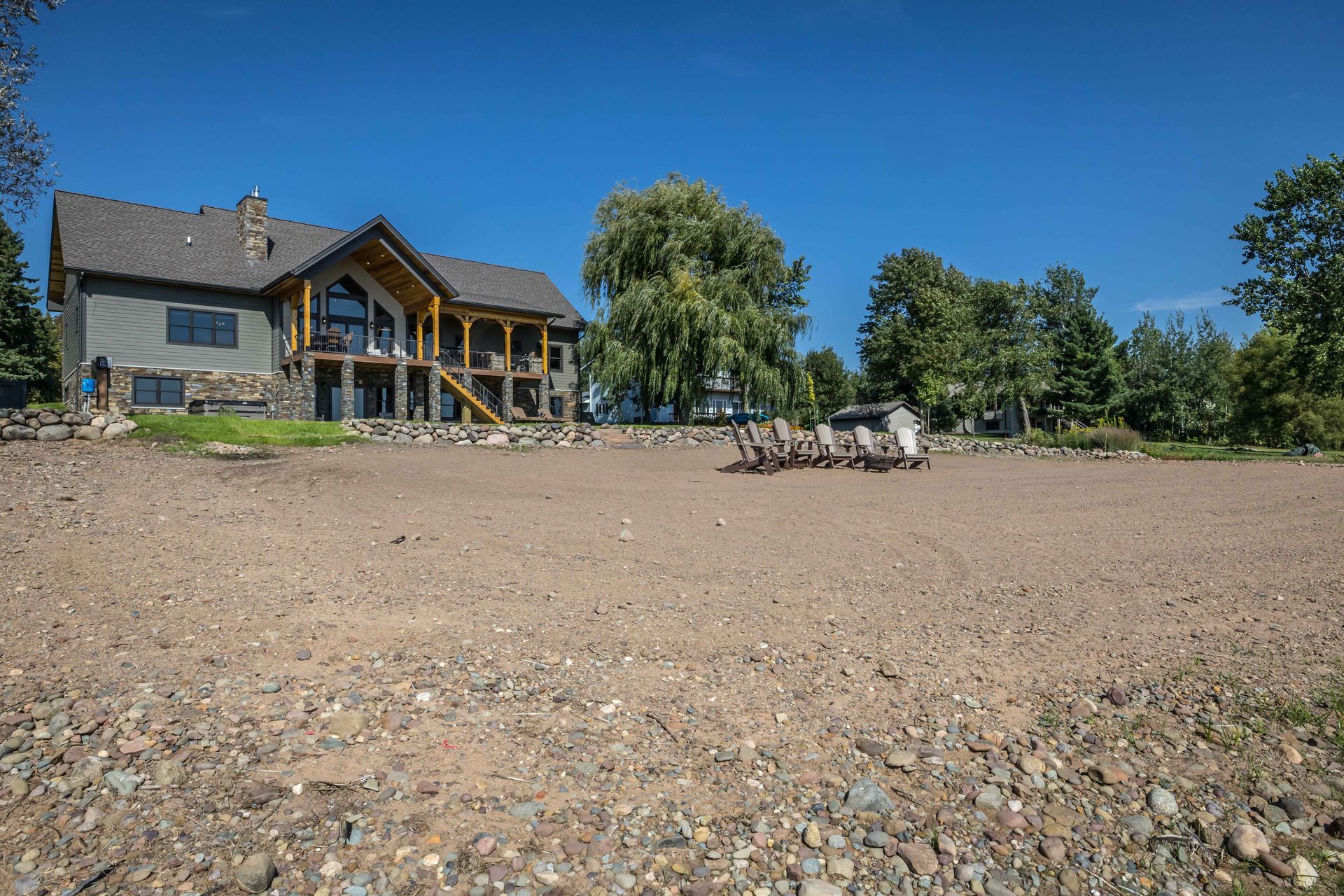 Large house with wood accents on a gravel shore under a blue sky, some trees, and a parked vehicle.