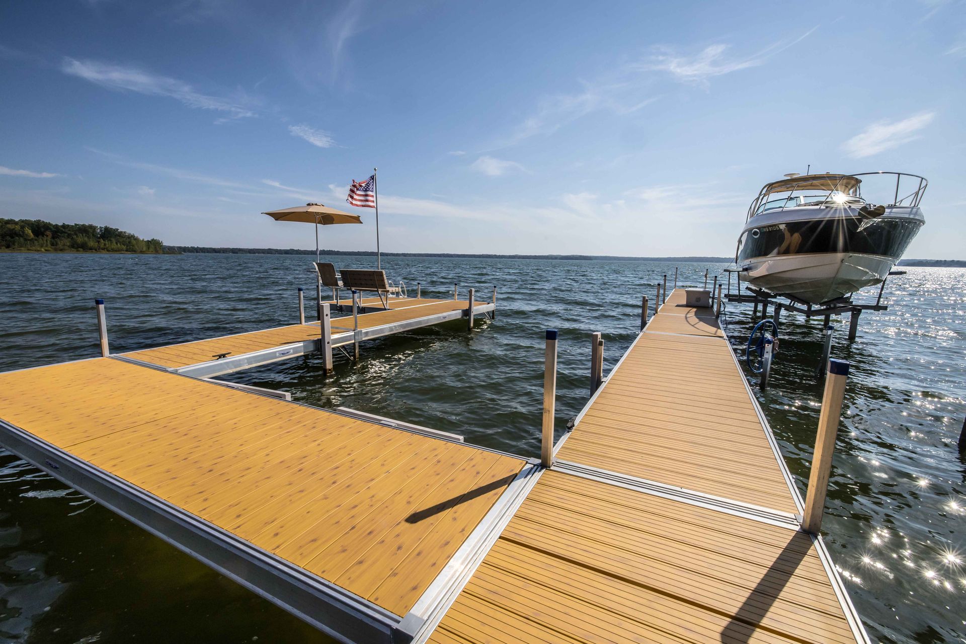 Dock on a lake with boat lift holding a boat, seating, American flag, and clear blue sky.