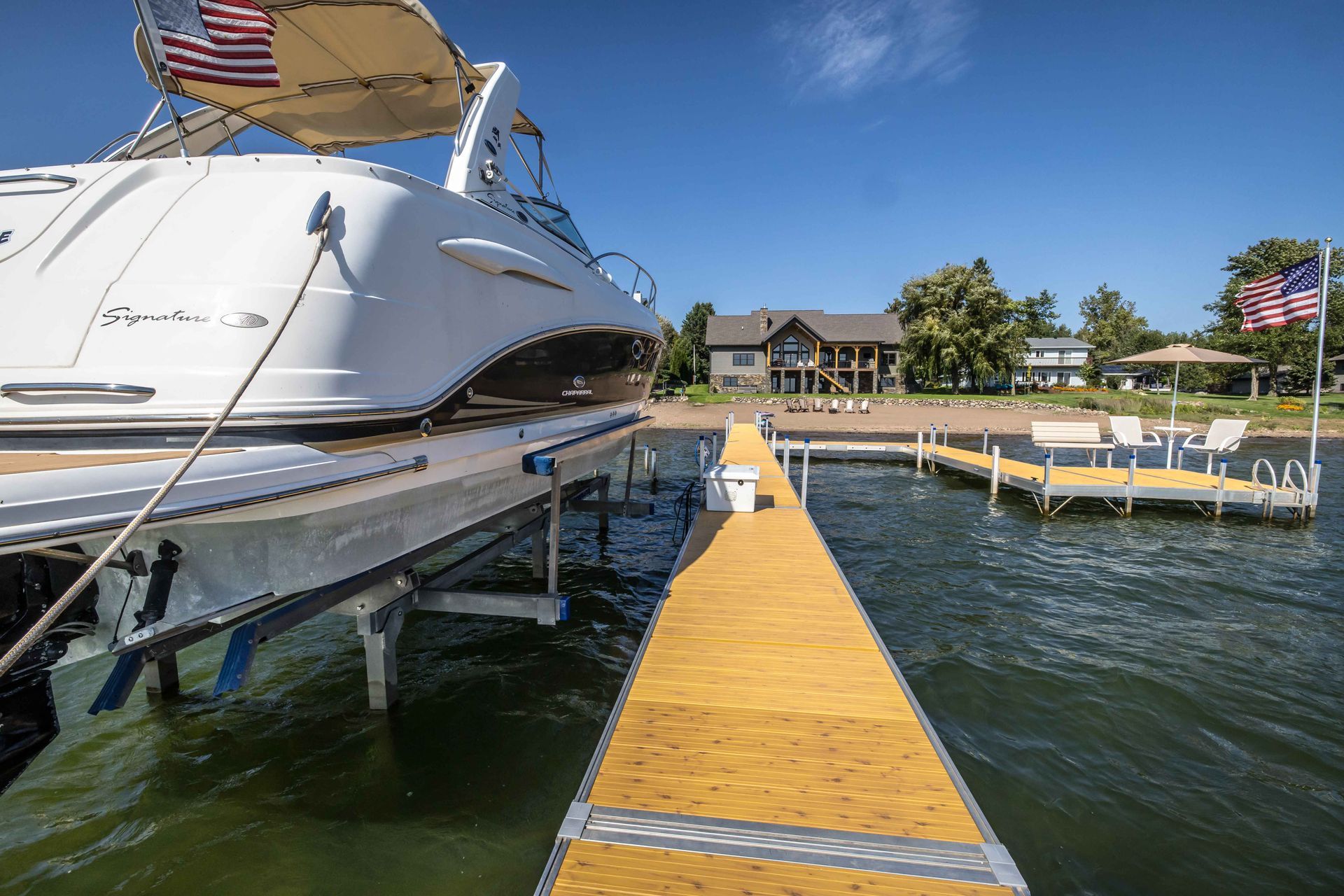 Boat docked on a lift at a wooden pier on a lake. Houses and beach in the background under a blue sky.