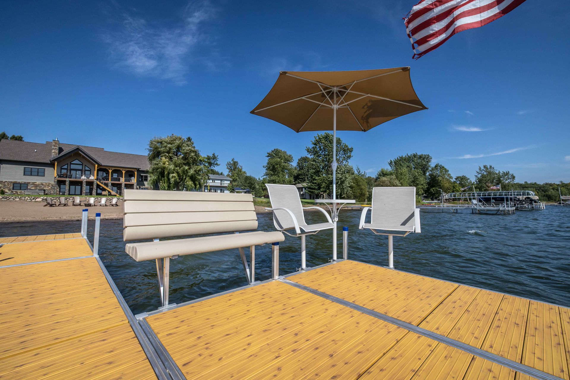 Dock with tan bench, chairs, umbrella, and American flag on a sunny day. Large house visible in the background.