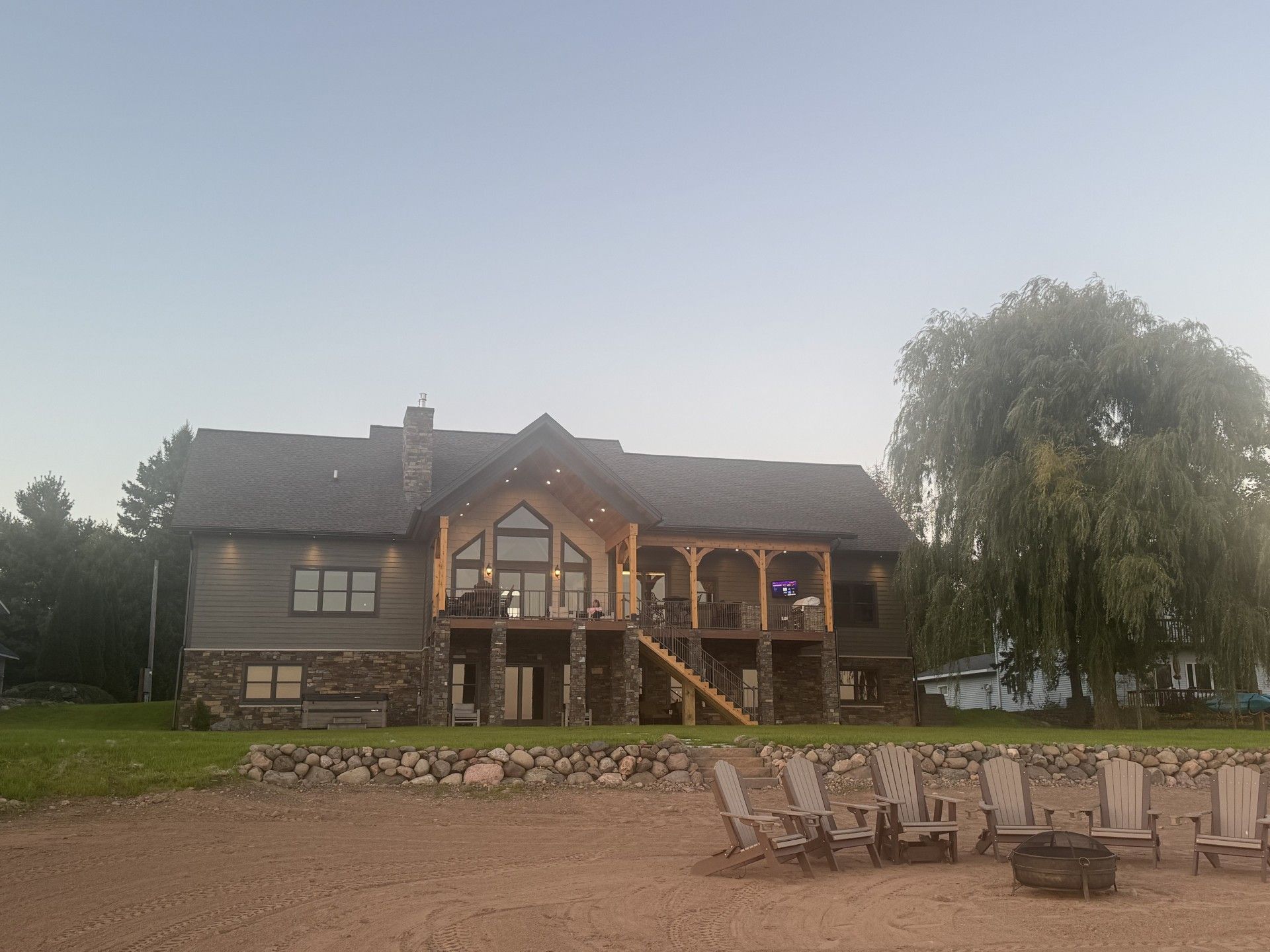 Large house on a beach with chairs; light brown siding, stone accents, wooden porch, and willow tree.