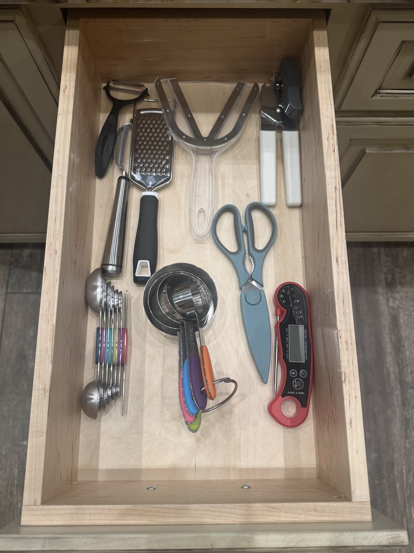 Kitchen drawer with various utensils: peeler, grater, measuring spoons, scissors, bottle opener, thermometer.