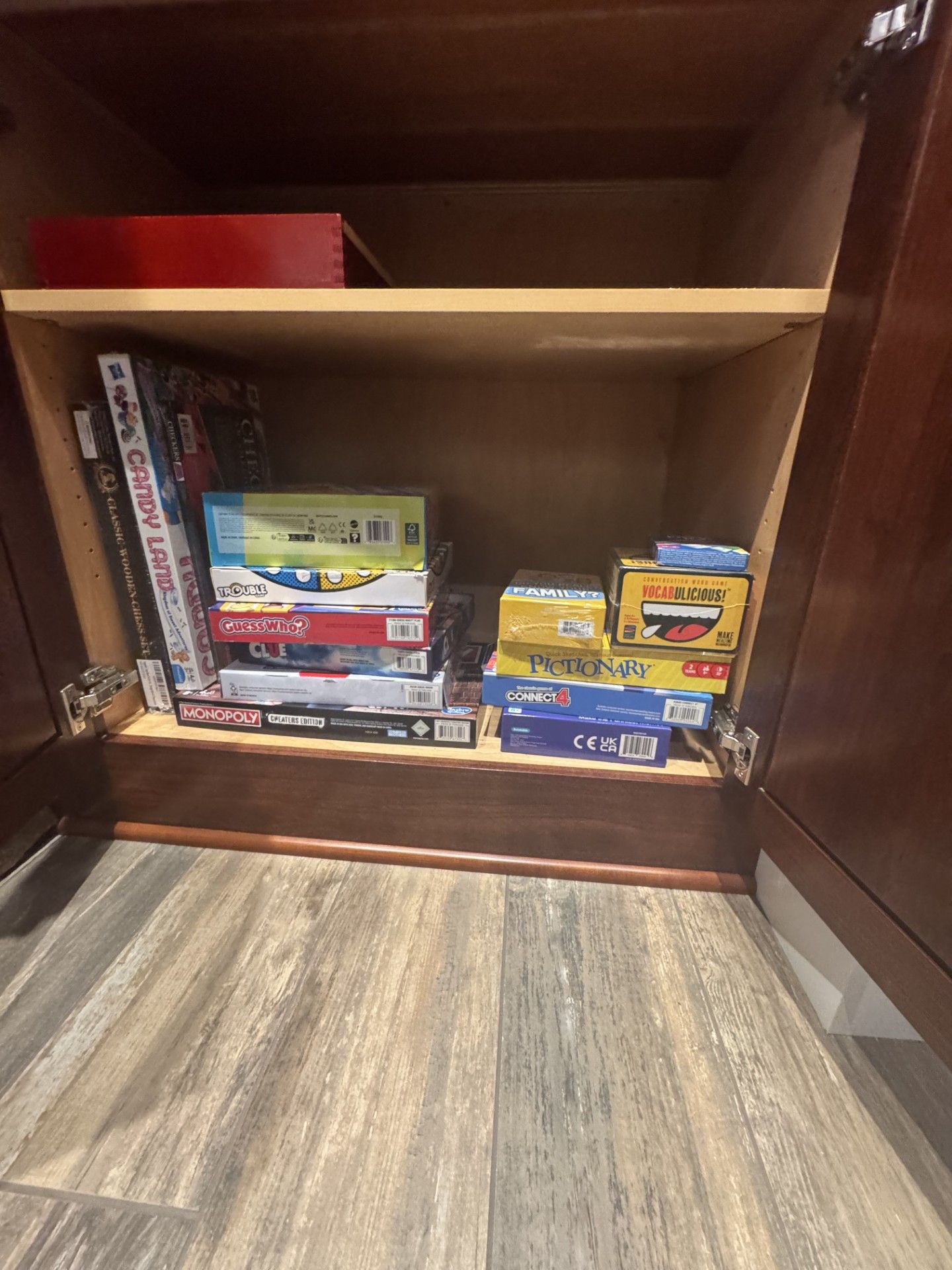 Wooden cabinet with games on a shelf. Floor is light wood, the cabinet is dark wood. Games are various colors.