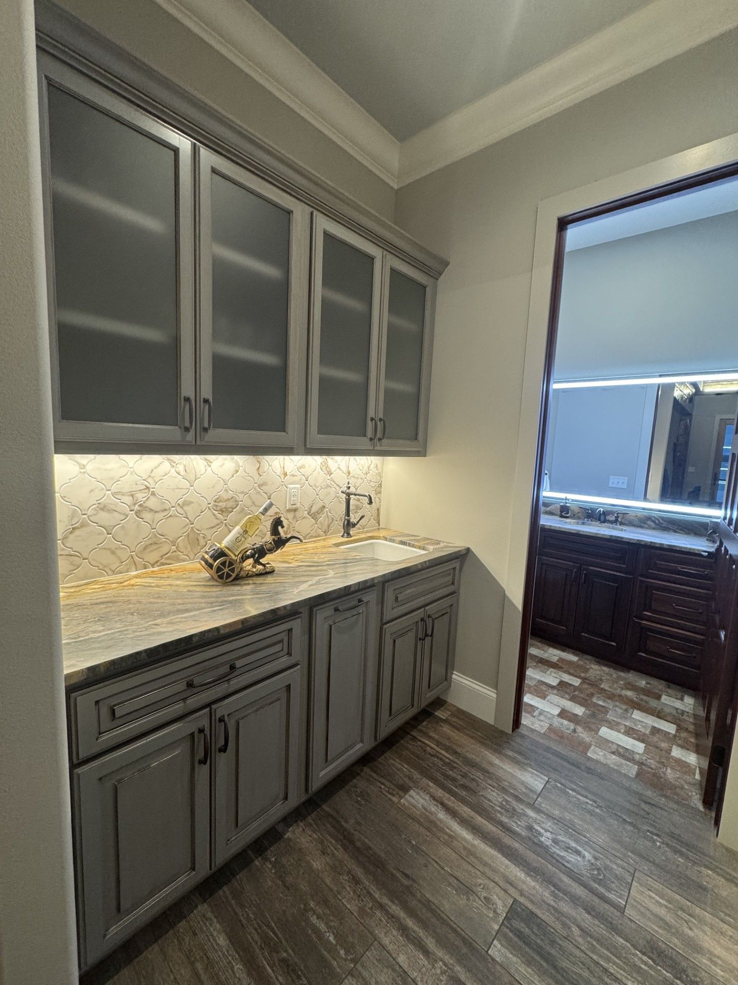 Gray kitchen cabinets with glass doors, countertop, and sink. Wooden floor, doorway to another room visible.