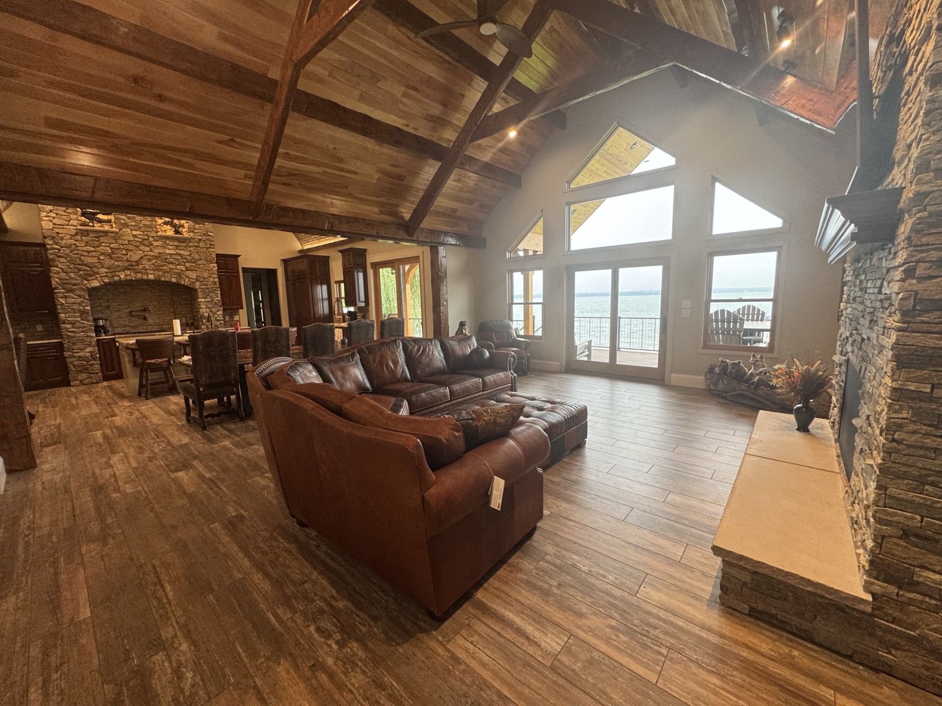 Spacious living room with wood floors, stone fireplace, leather sofa, and large windows overlooking a lake.