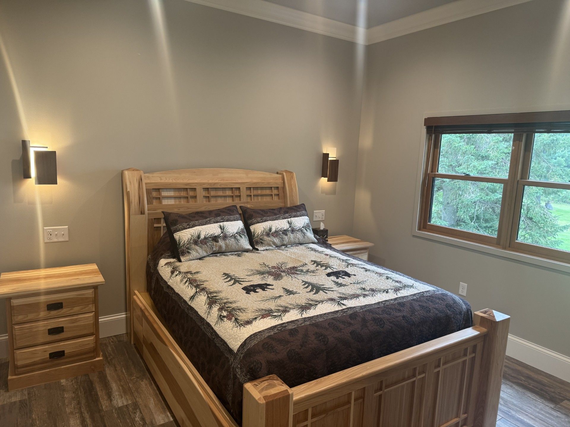 Bedroom with a light wood bed, nightstands, and a patterned quilt. Wall sconces and a window are present.