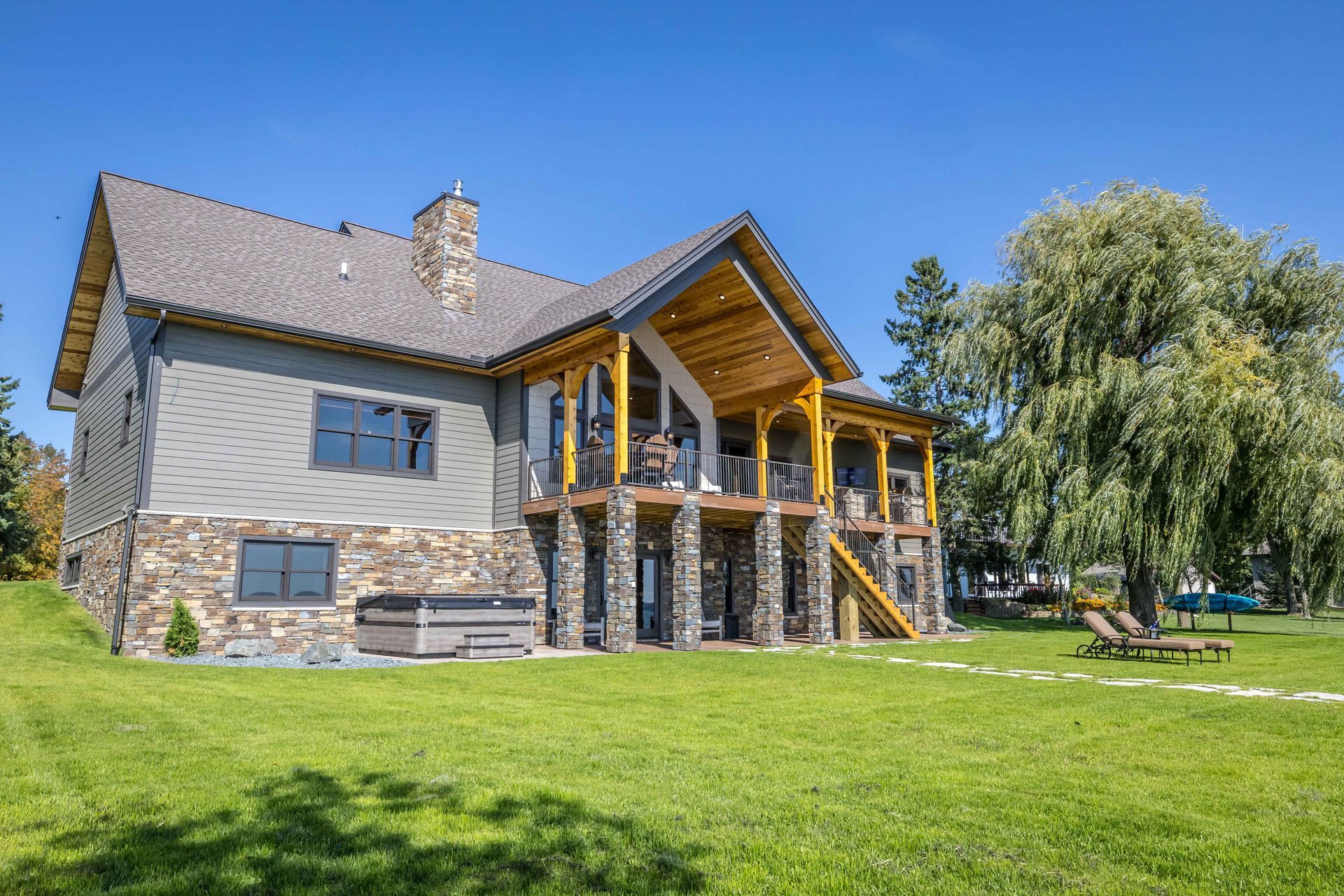 Lakefront home with stone facade, balcony, hot tub, and lush green lawn under a blue sky.