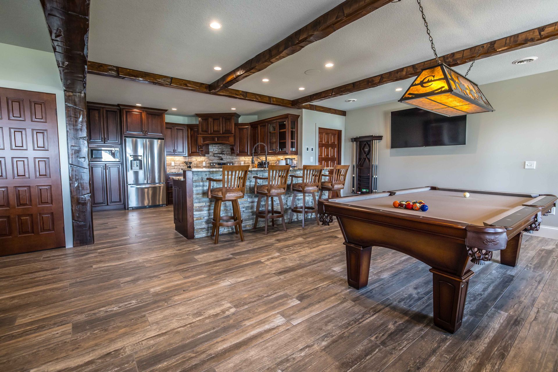 Game room with a pool table, kitchen, and bar. Brown wooden flooring and cabinetry.