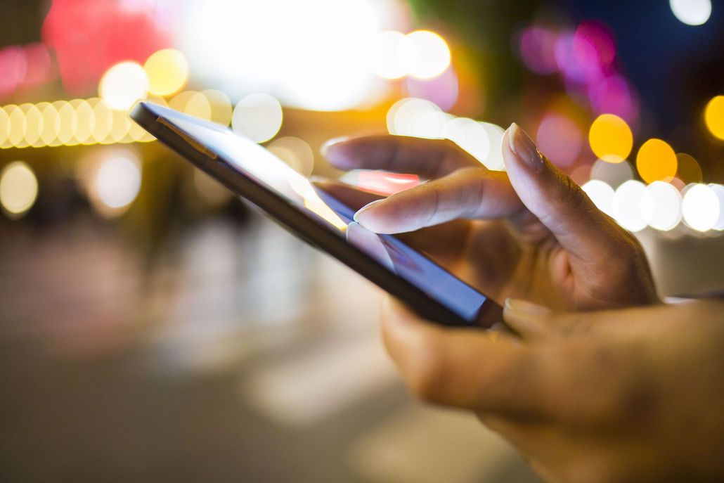 Hand holding a smartphone, fingers touching the screen. Blurred city lights in the background.