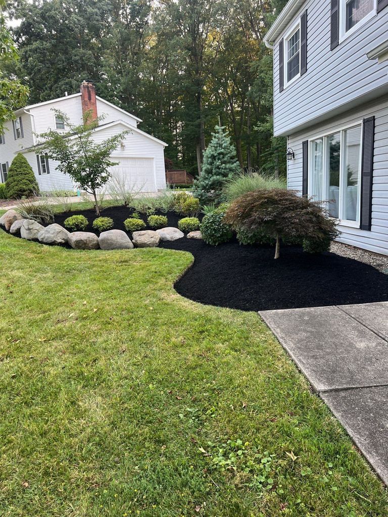 A house with gray siding and black shutters, surrounded by a lawn, garden, and trees. - After