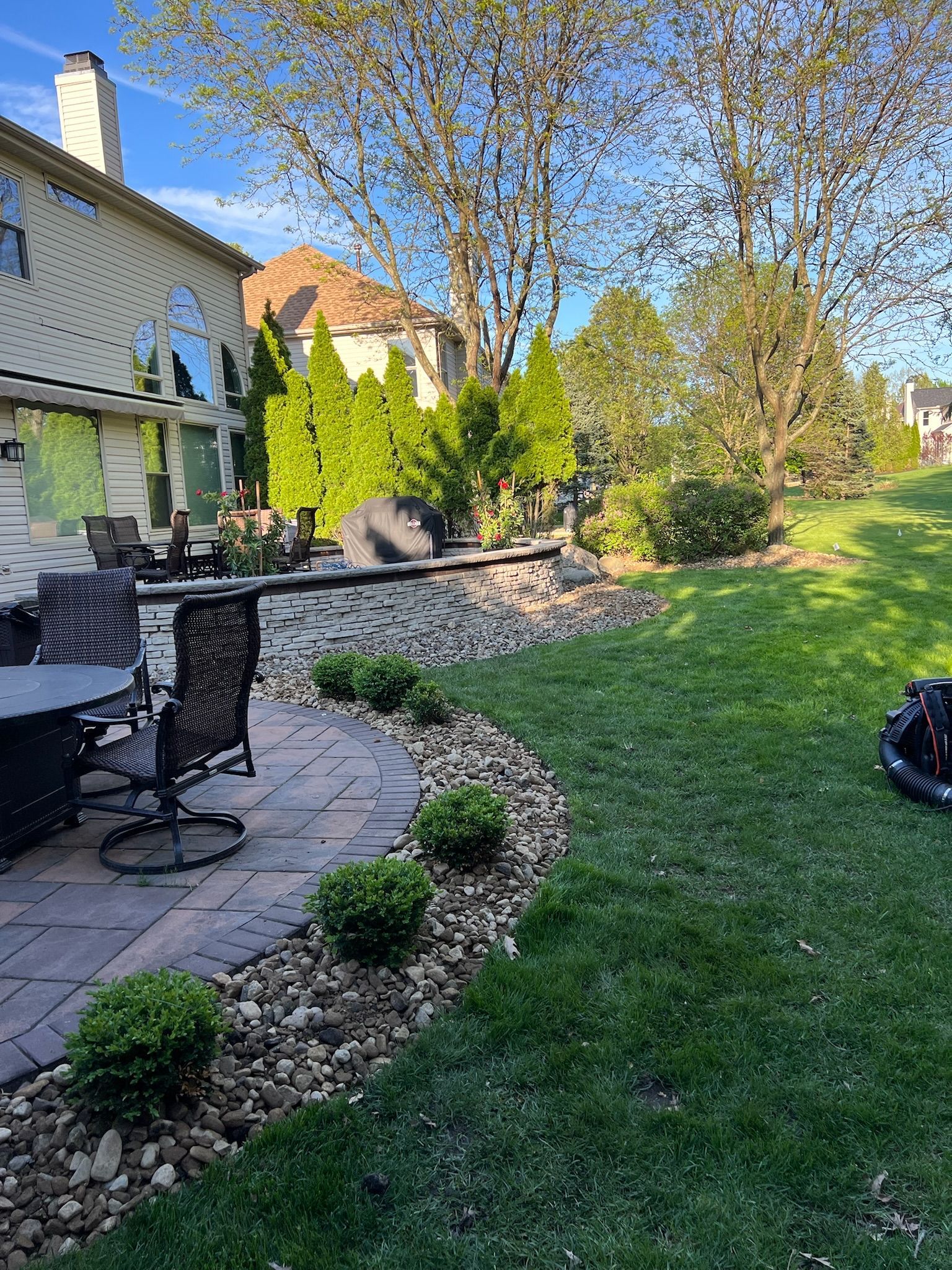 Patio with chairs, shrubs, and a stone border, adjacent to a lawn and a house on a sunny day.