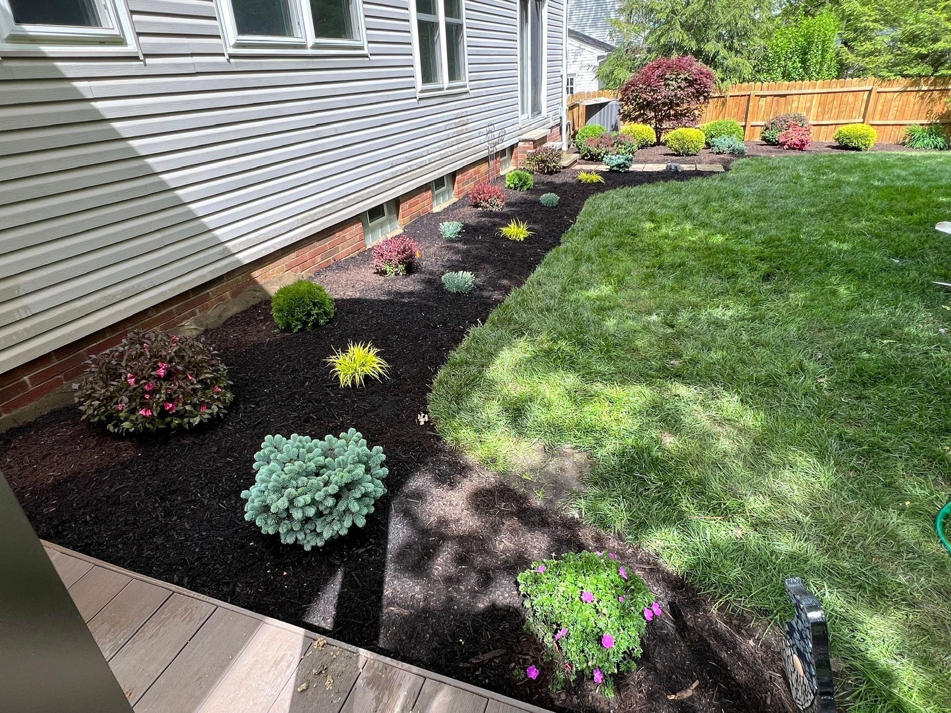 Landscaped garden bed with dark mulch and various colorful plants next to a house and lawn.
