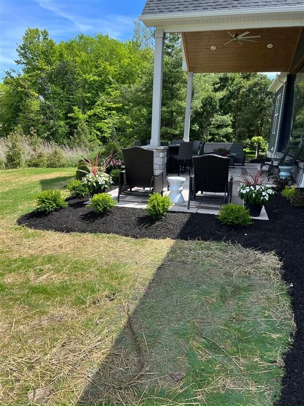 Patio with black mulch, green bushes, chairs, and plants, next to a grassy lawn on a sunny day.