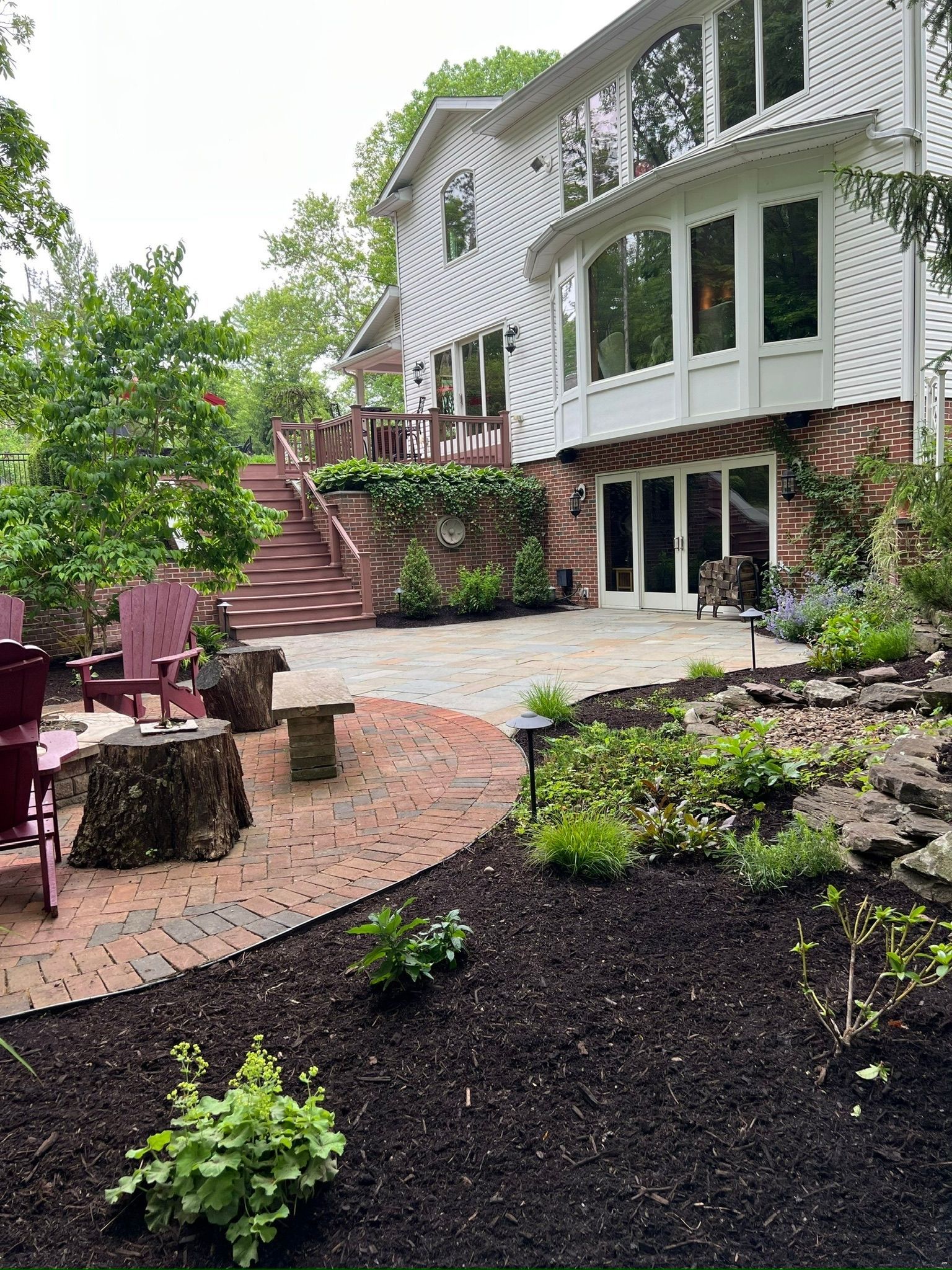 A two-story white house with a stone patio and steps leading up to a deck; landscaping with red brick and mulch.