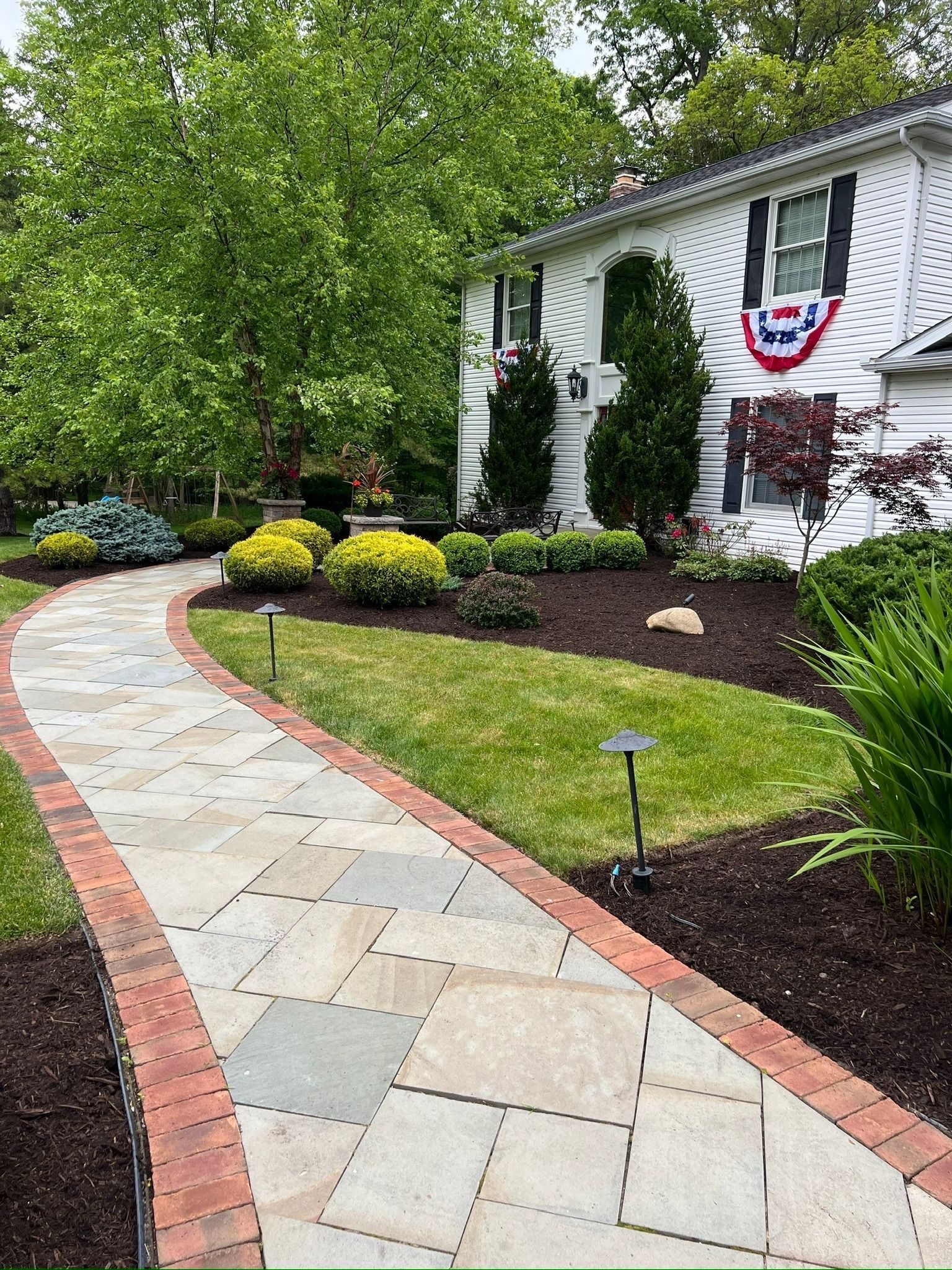 A winding stone walkway bordered with brick leads to a white house with a decorative garland.