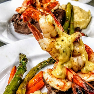 Grilled shrimp with sauce, steak, asparagus, carrots, and mashed potatoes on white plates.
