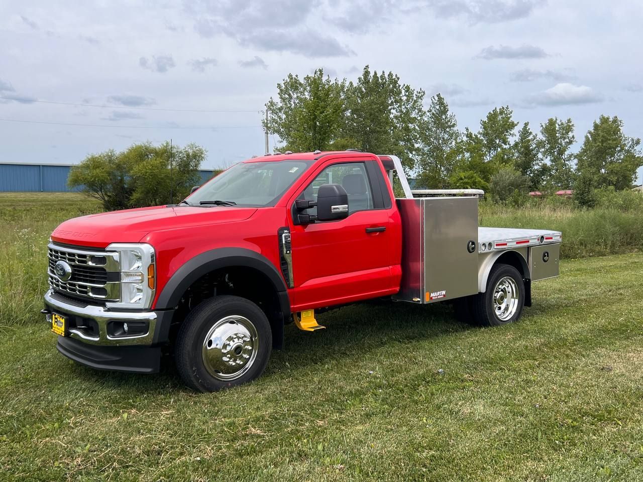Red Ford truck with a flatbed in a grassy field. The truck is new, with chrome wheels and a tool box.