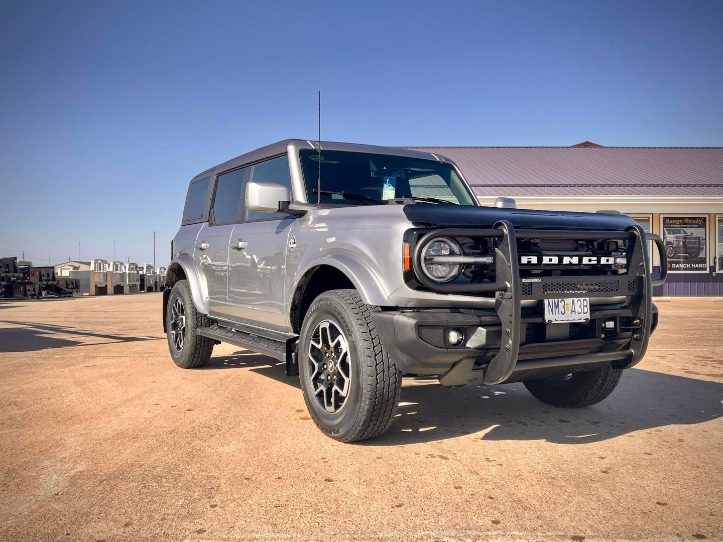 Silver Ford Bronco SUV parked on dirt, with a black brush guard, in front of a building on a sunny day.