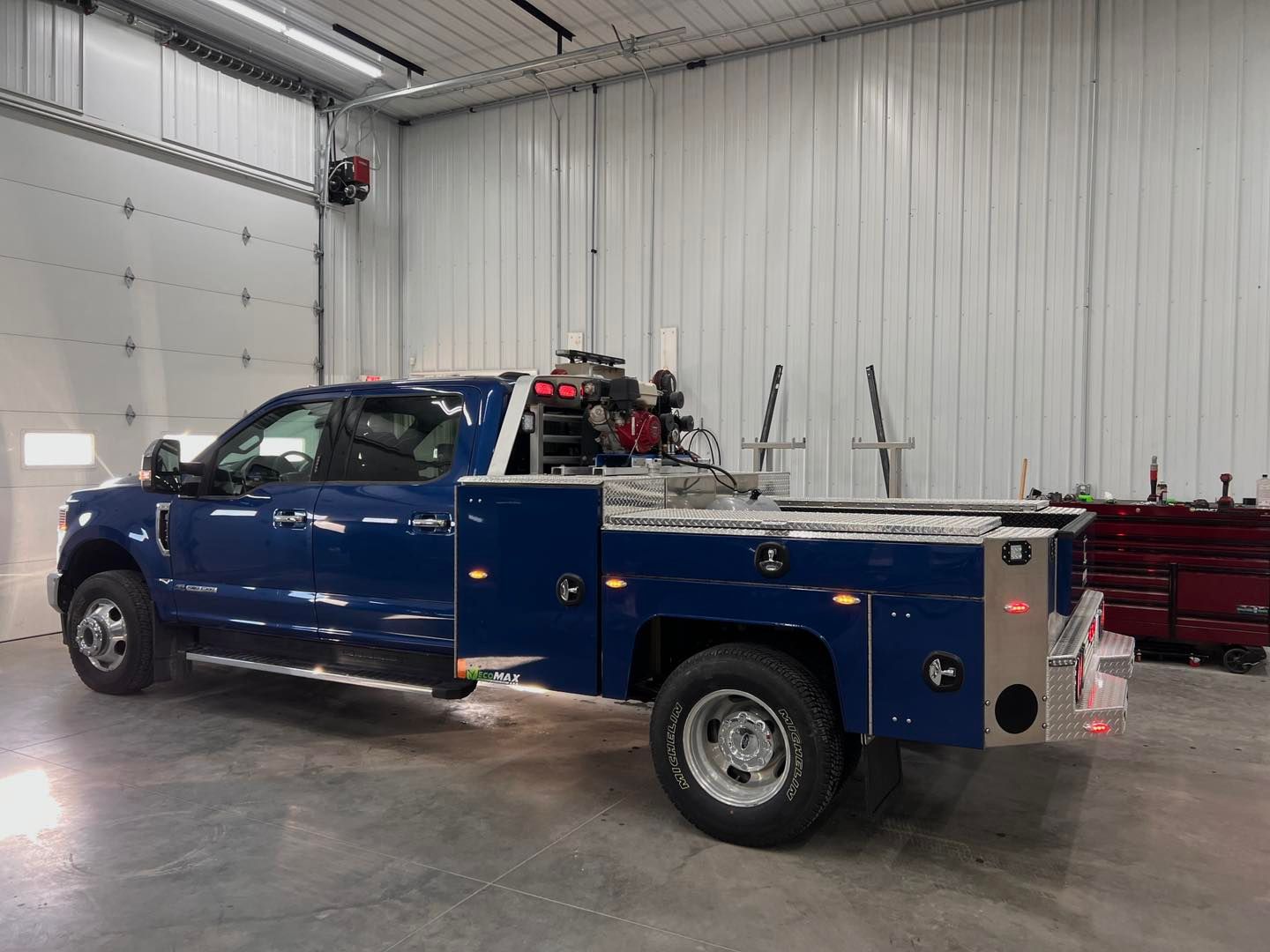 Blue work truck parked inside a garage. The truck has a flatbed with tool compartments.