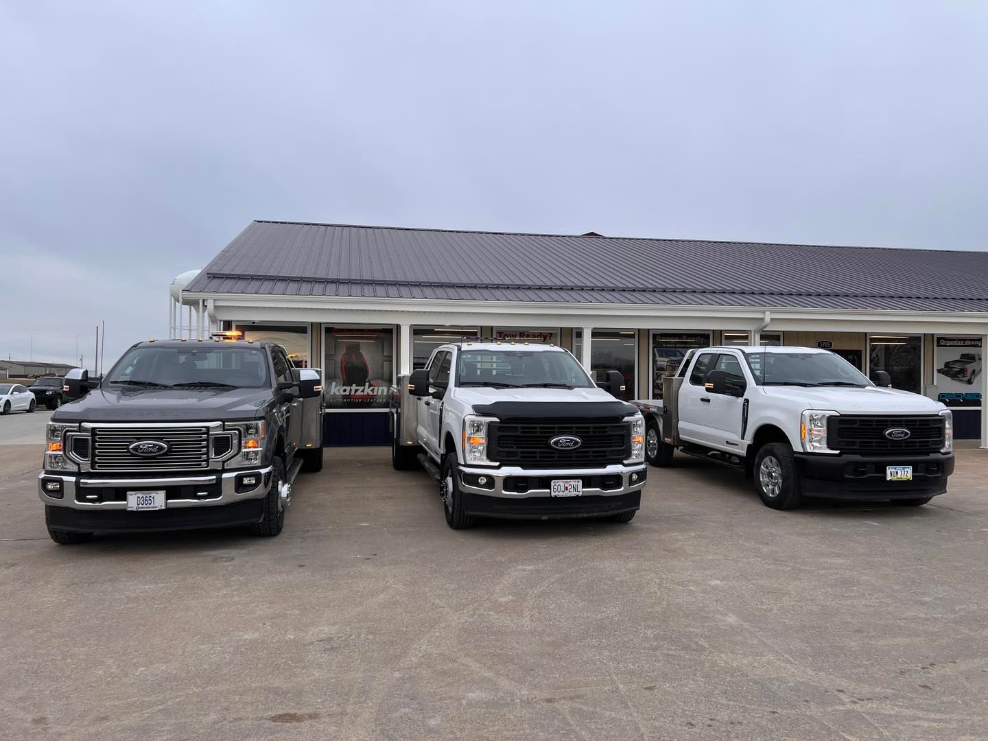 Three Ford pickup trucks parked in front of a building with windows, on an overcast day. Trucks are gray and white.
