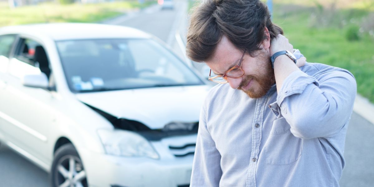 Man clutching neck in pain near a damaged white car, possibly a car accident.