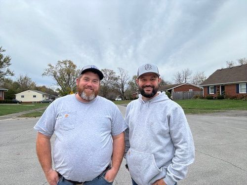 Two men standing on a street, smiling in gray hoodies and caps under a cloudy sky