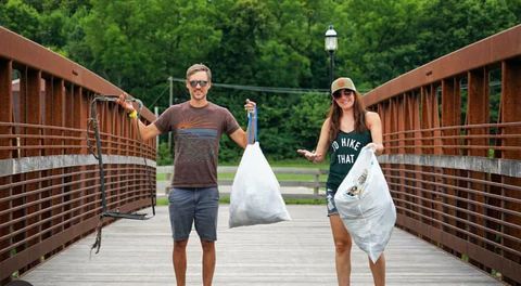 A man and a woman are standing on a bridge holding bags of trash.