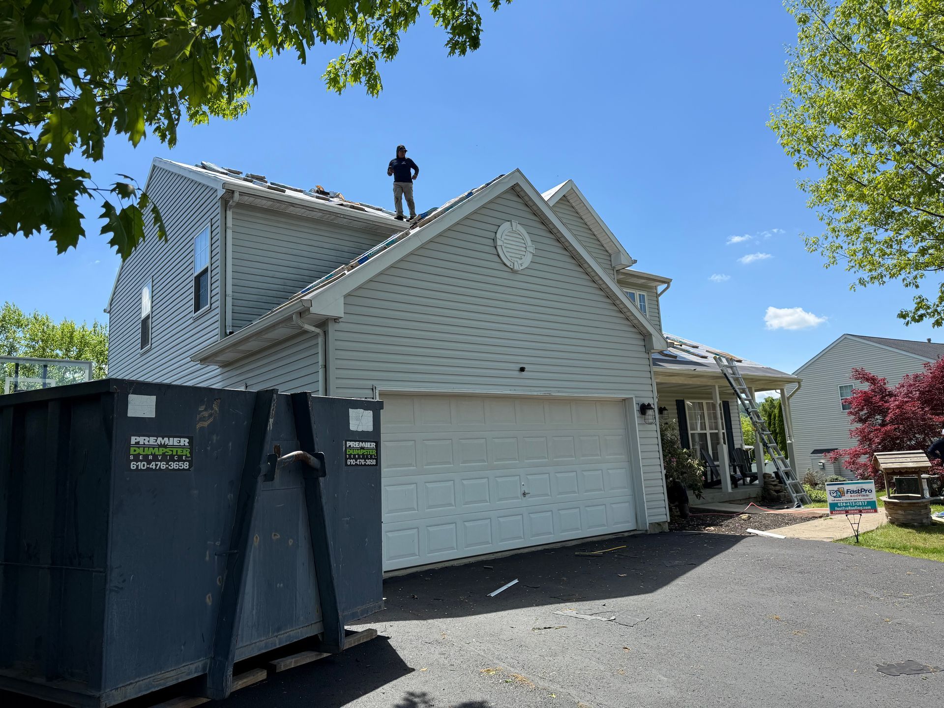 A man is standing on the roof of a house next to a dumpster
