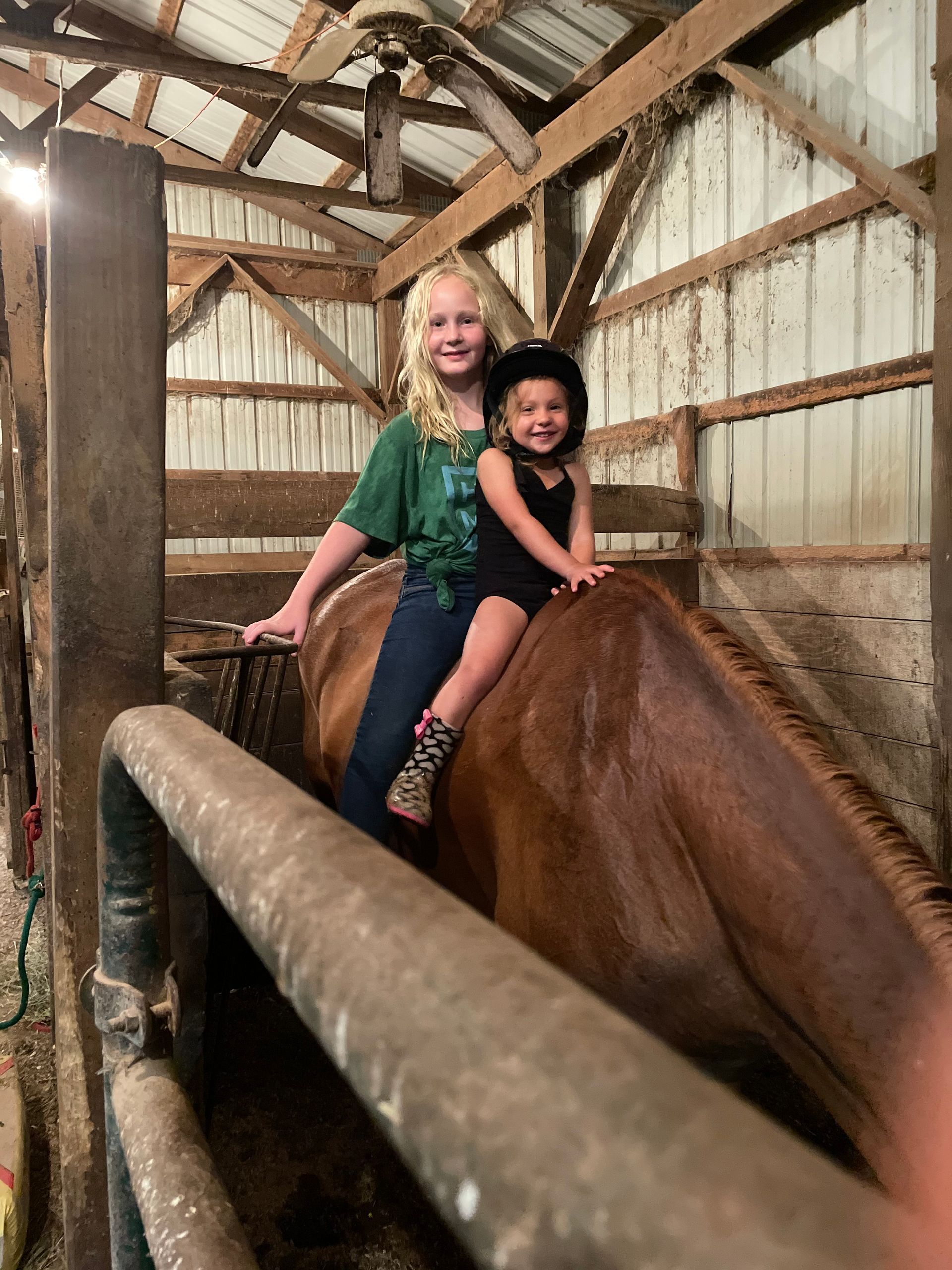 Two young girls are riding on the back of a brown horse in a barn.