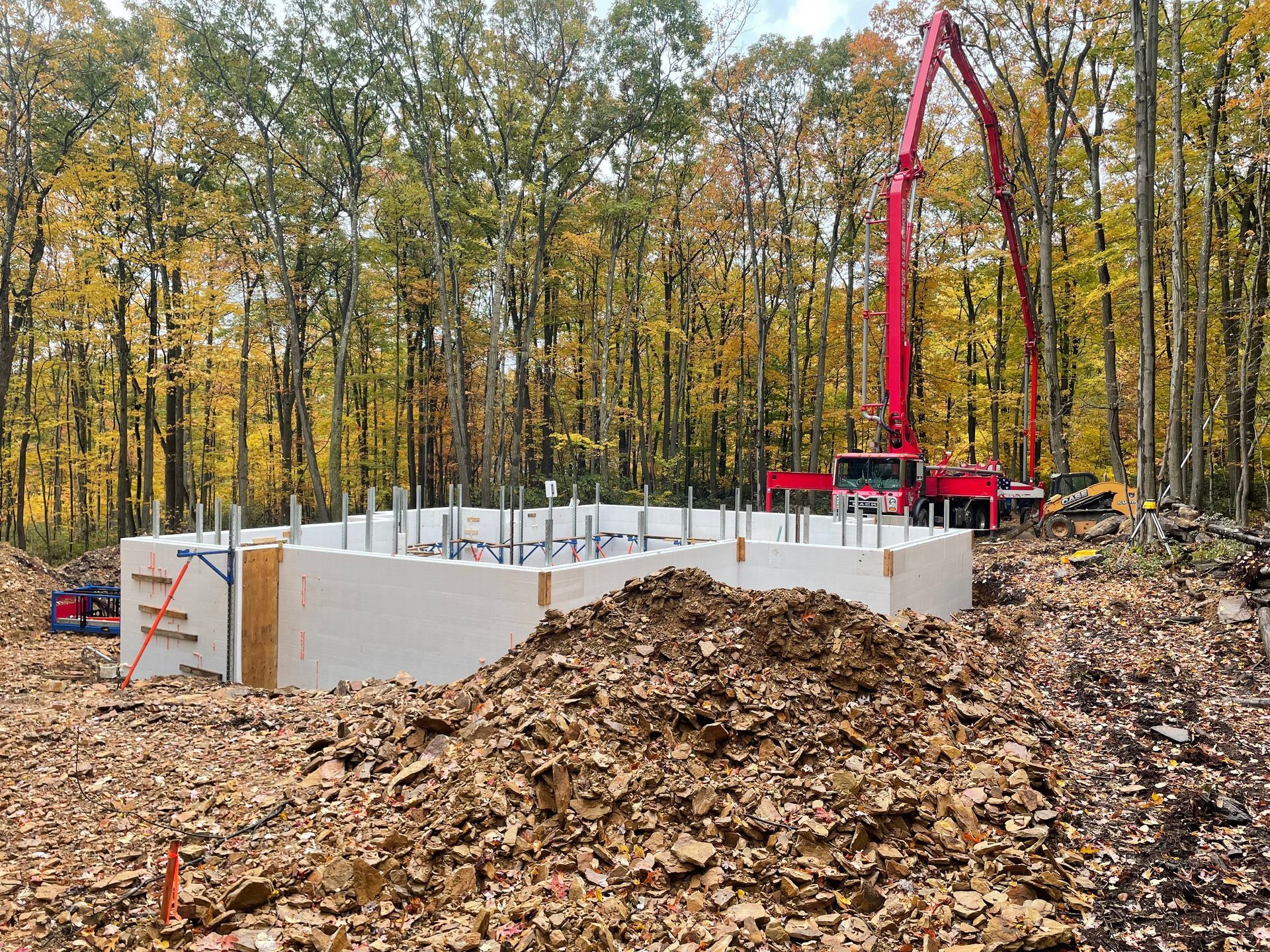 A red crane is pumping concrete into a foundation in the middle of a forest.