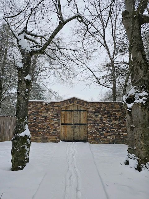 A wooden gate is surrounded by snow covered trees and a stone wall.
