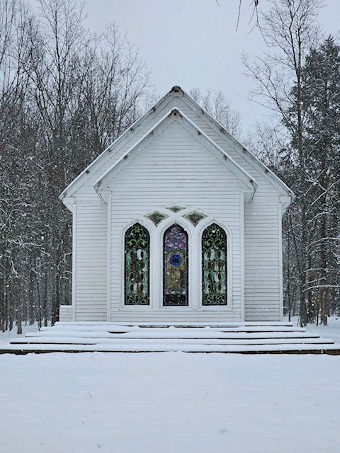 A small white church with stained glass windows is surrounded by snow.