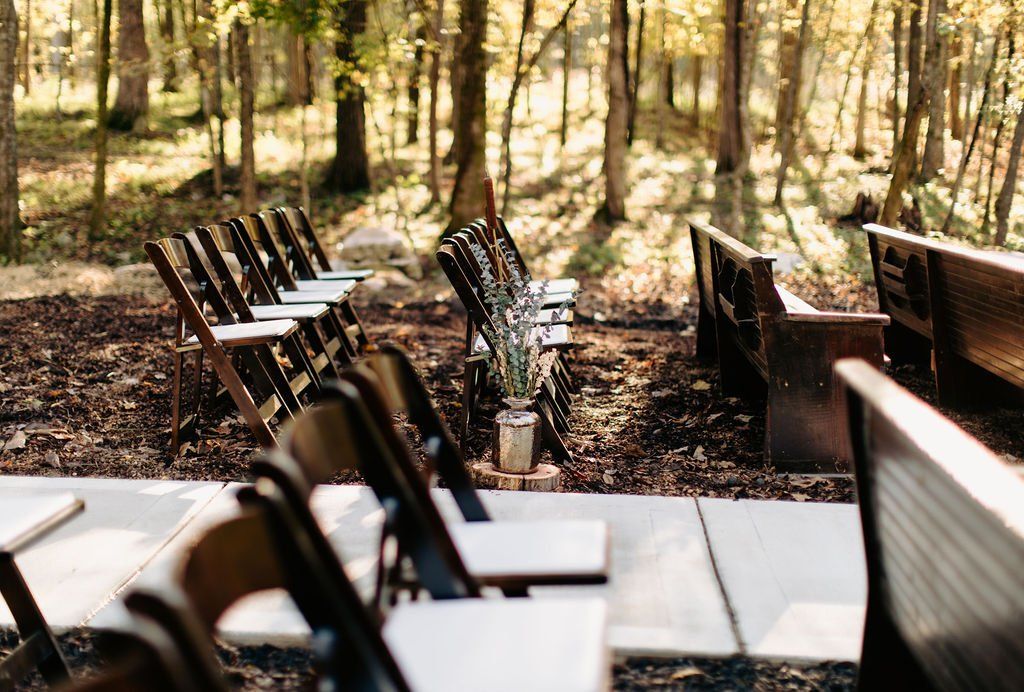 A row of folding chairs are lined up in the woods for a wedding ceremony.