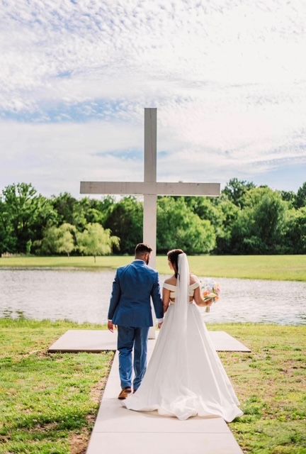 Bride and groom walking toward a large cross by a river, with a green field and cloudy sky behind them.