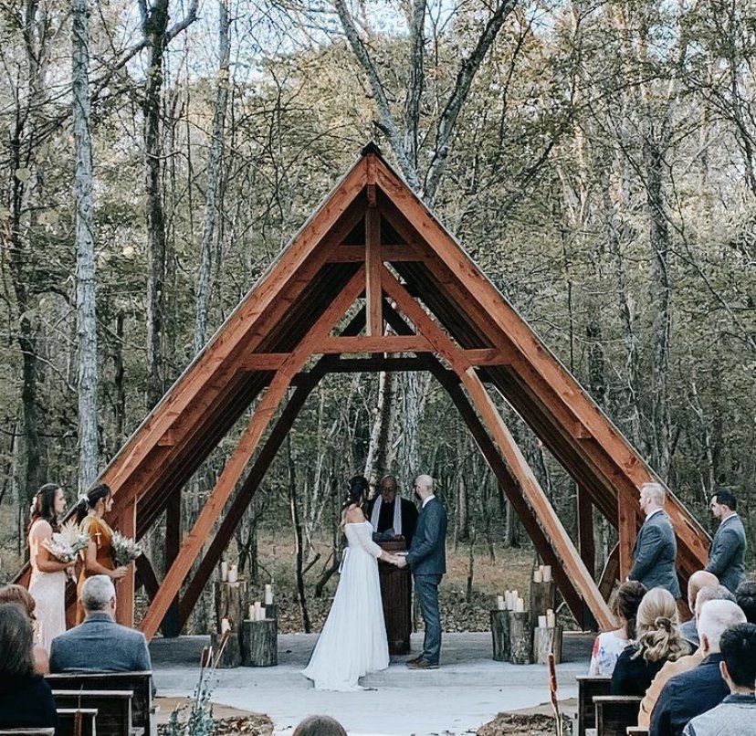 A bride and groom are getting married under a wooden structure in the woods.
