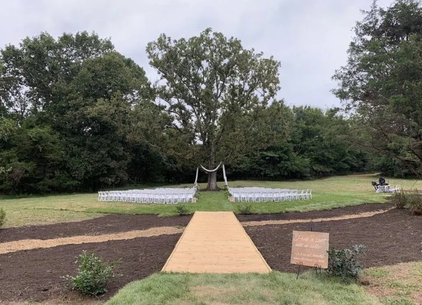 A wooden walkway leading to a wedding ceremony in a field surrounded by trees.