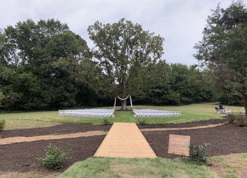 A wooden walkway leading to a wedding ceremony in a field surrounded by trees.