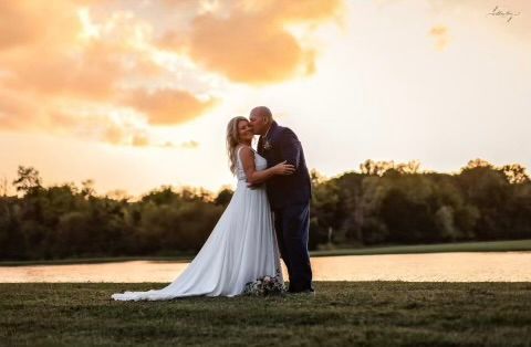 A bride and groom are kissing in front of a lake at sunset.