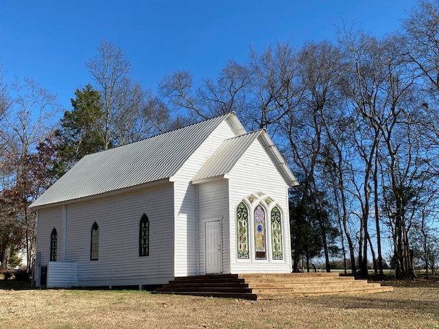 A small white church with stained glass windows is surrounded by trees.