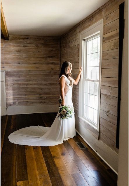A bride in a long white dress is standing in front of a window.