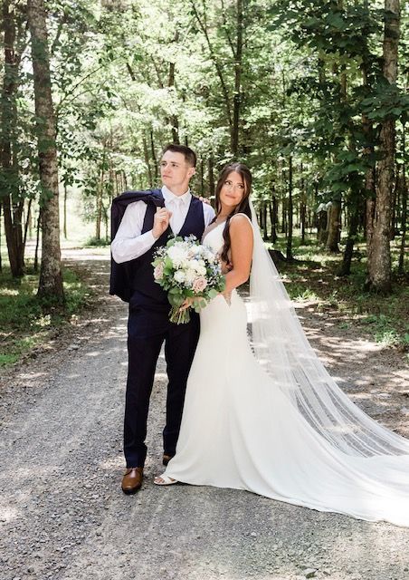 A bride and groom are standing next to each other on a dirt road in the woods.