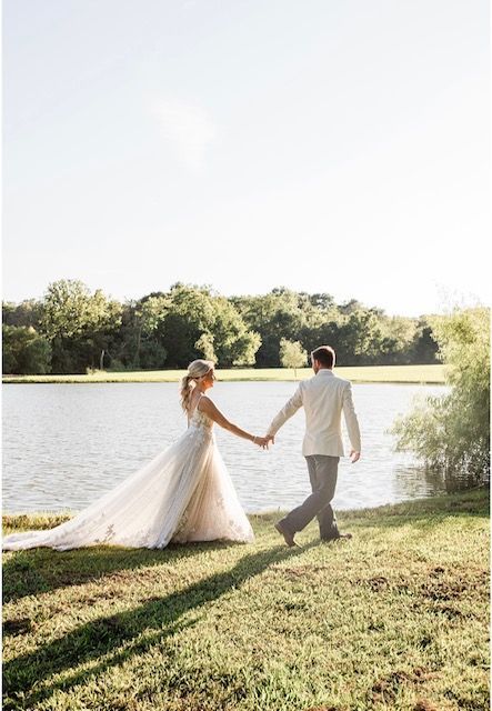 A bride and groom are walking by a lake holding hands.