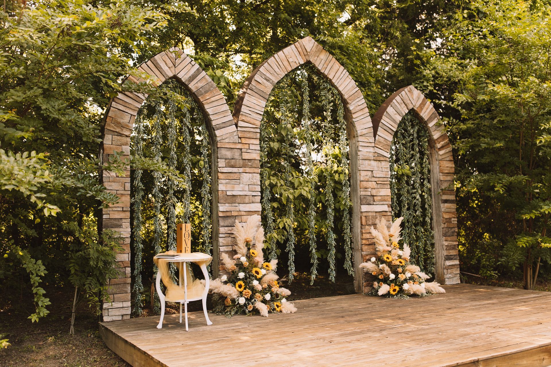 A wooden stage with arches and flowers on it in the middle of a forest.