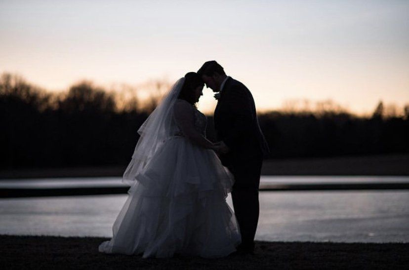 A bride and groom are kissing in front of a lake at sunset