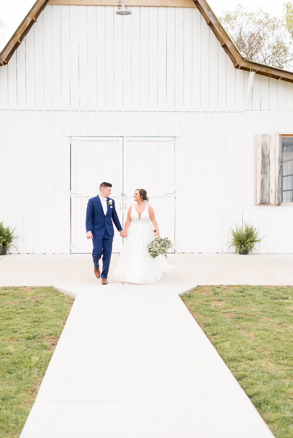 A bride and groom are walking down a path in front of a white barn.