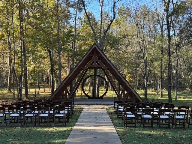 Outdoor wedding ceremony setup with rows of chairs and a wooden triangular arch in a wooded clearing.