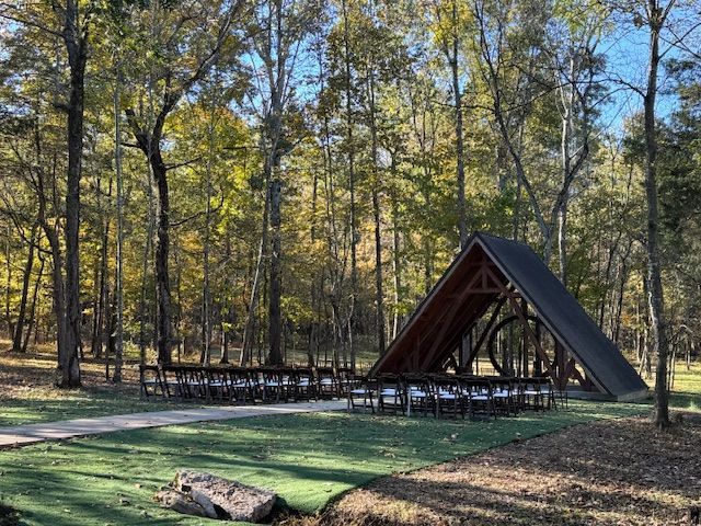 A-frame cabin in a wooded park with a picnic table and path under tall trees.