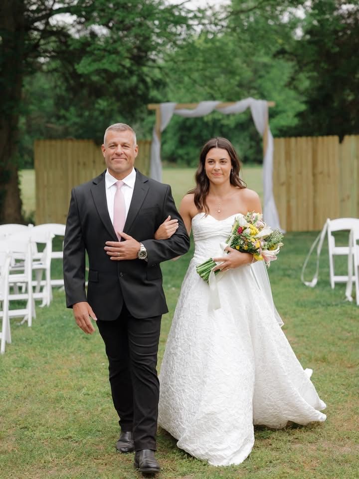 Bride and suited escort walking down the aisle outdoors, with white chairs and greenery in the background
