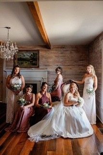 Six women in formal dresses pose in a rustic room, with the bride seated in a white gown holding a bouquet.