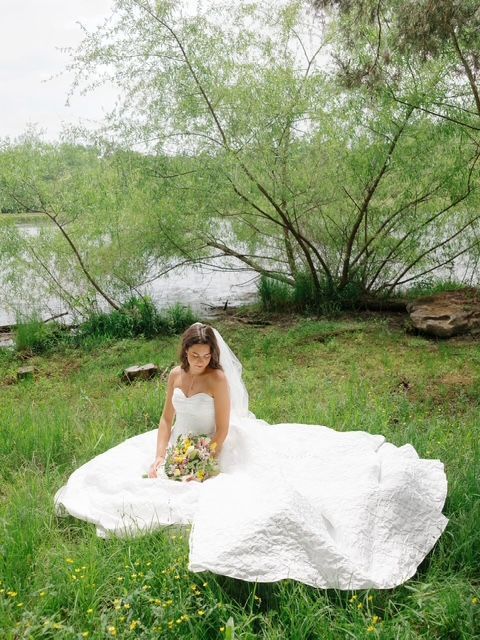 Bride in a white gown sitting on grass by a tree, holding a bouquet outdoors by the water.